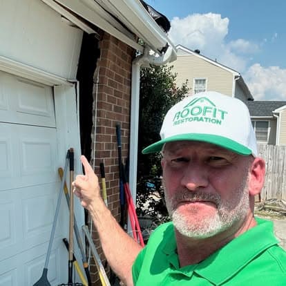Man in green shirt and cap pointing at home damage near garage entrance. RoofFit Restoration.