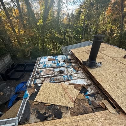 Damaged roof with exposed plywood and chimney, surrounded by autumn foliage.