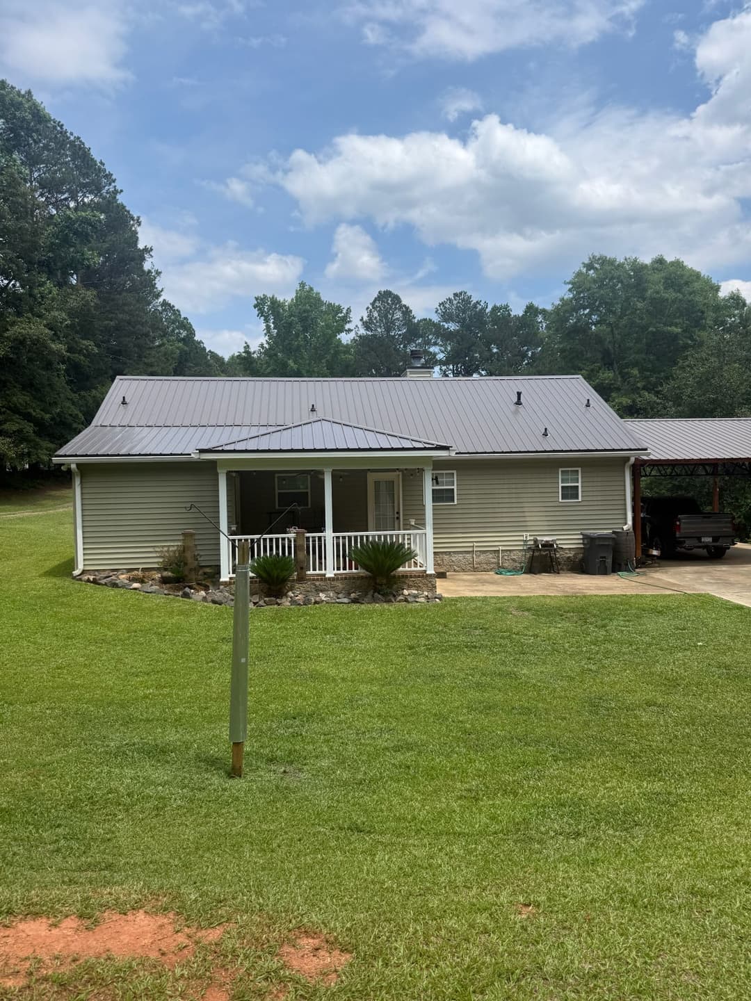 Single-story home with a metal roof, green lawn, and surrounding trees on a sunny day.