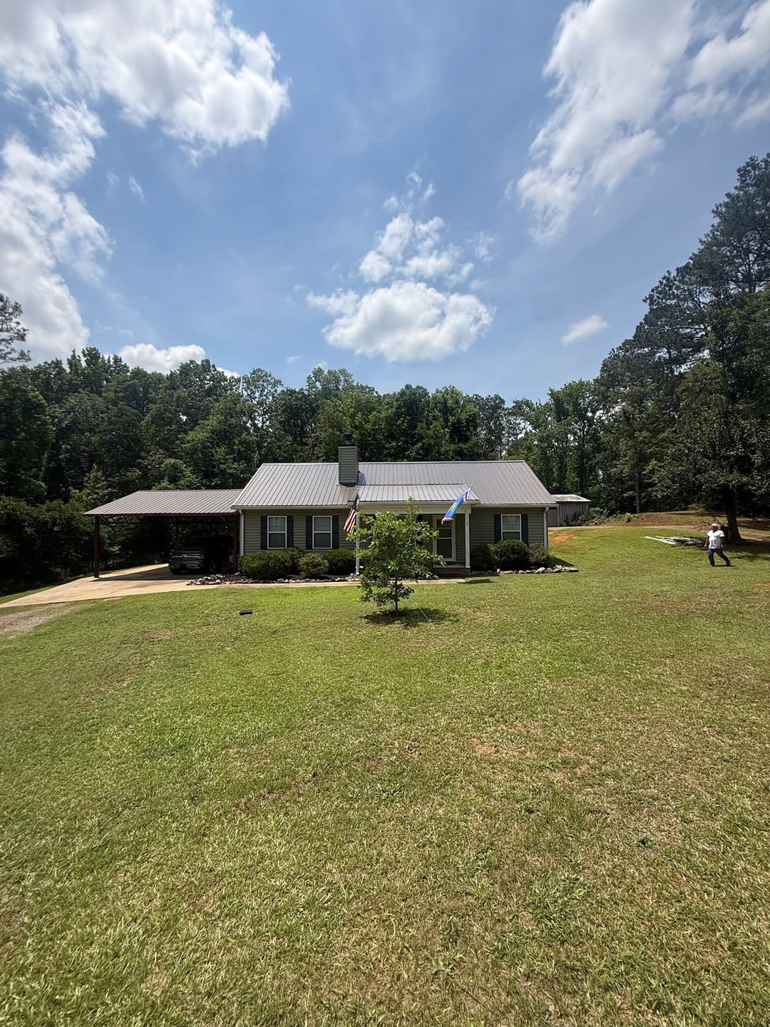 Single-story house with a metal roof, surrounded by trees and a grassy lawn on a sunny day.