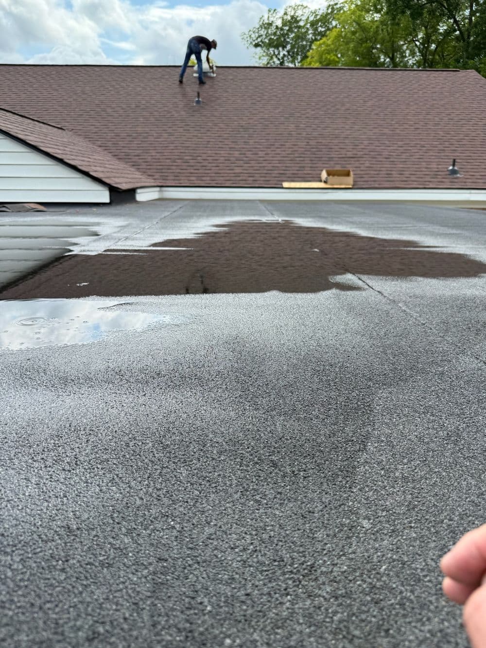 Roofer applying shingles on a sloped roof with puddles on a flat roofing surface.