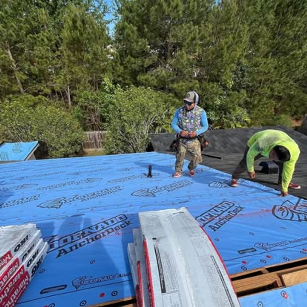 Two workers installing roofing materials on a residential roof with trees in the background.